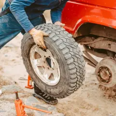 Persona con guanti smonta una gomma auto da un fuoristrada rosso. Si vede un cric arancione a terra, utile per capire dove smaltire gomme auto usate.