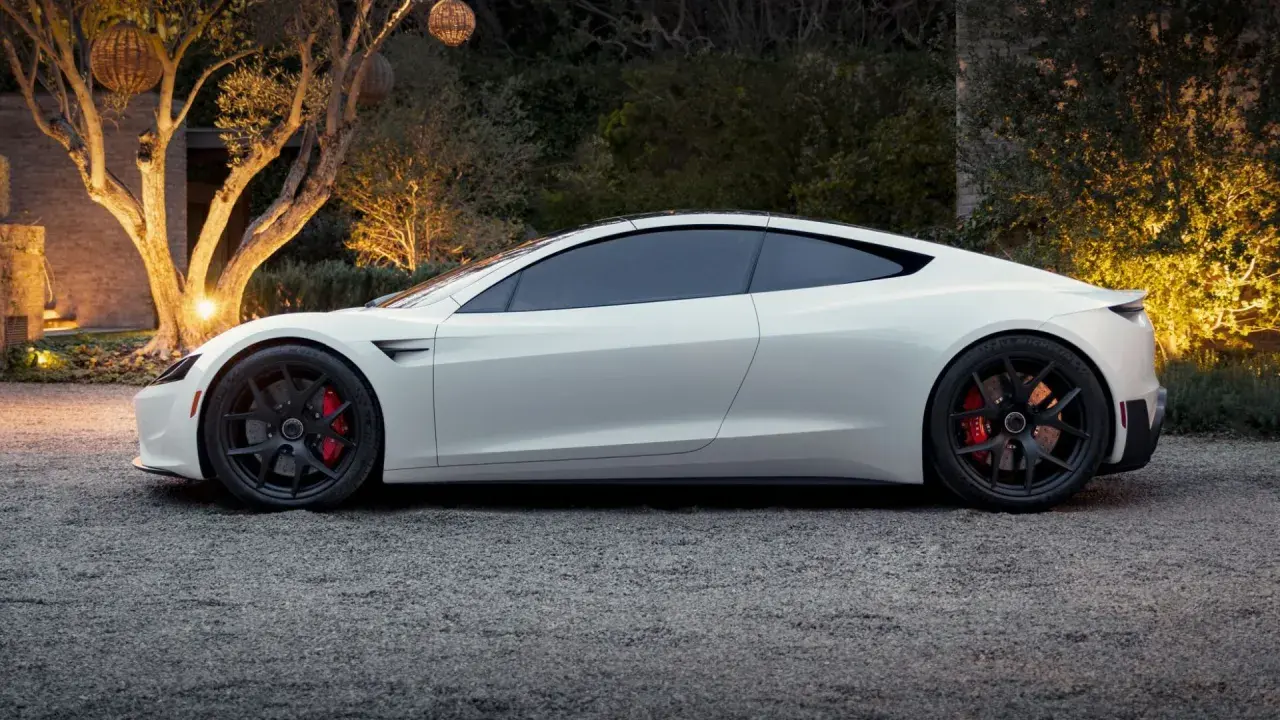 Bianca Tesla Roadster parcheggiata su ghiaia, con luci soffuse che illuminano alberi e vegetazione sullo sfondo.