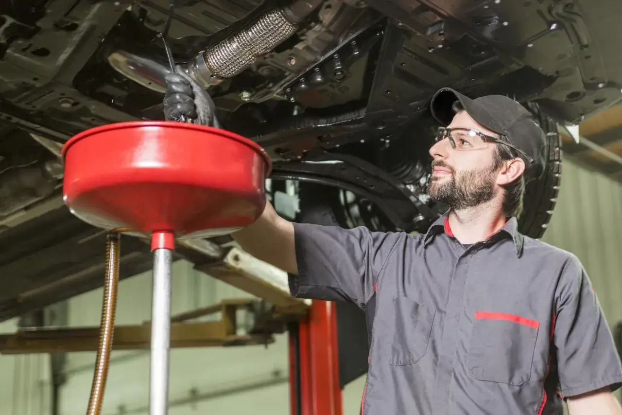 Meccanico con guanti e occhiali lavora sotto un'auto, raccogliendo liquidi in un contenitore rosso. Un passo essenziale per la decarbonizzazione motore.